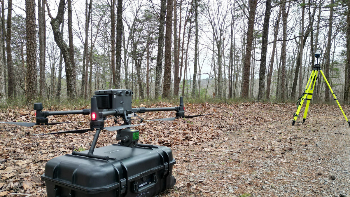 Survey drone on its equipment case beside a tripod-mounted receiver in a wooded field location.
