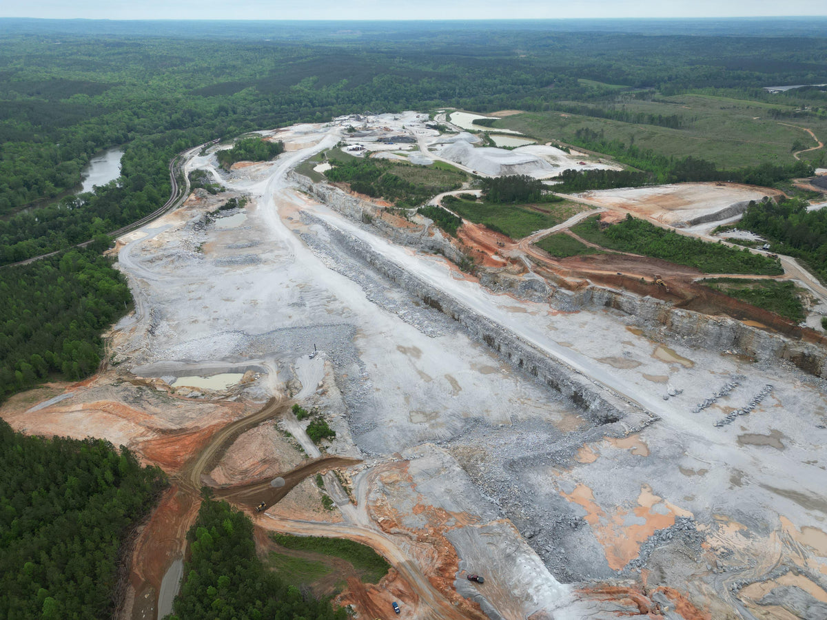 Aerial view of a quarry with terraced rock faces, access roads, stockpiles, a nearby river, and surrounding forest.