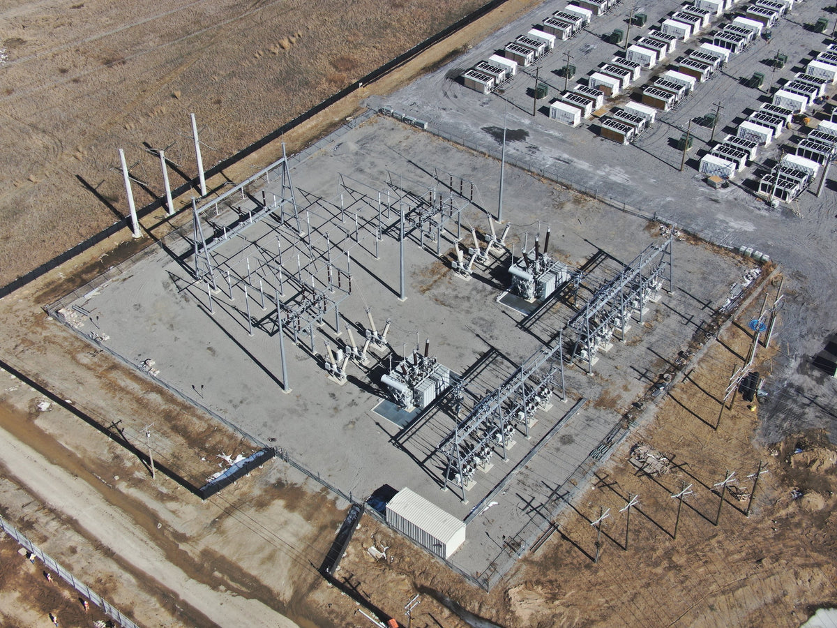 Aerial view of an electrical substation with transformers, steel structures, and rows of industrial cooling equipment behind it.