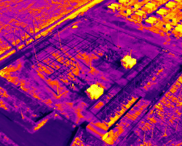 Aerial infrared image of an electrical substation showing transformers, electrical structures, and adjacent industrial cooling equipment in thermal false colors.